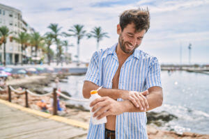 Male smiling while applying sunscreen lotion at the beach for sun protection.